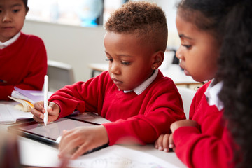 Three kindergarten school kids sitting at desk in a classroom using a tablet computer and stylus together, selective focus