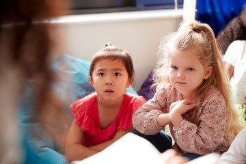 Two infant school girls sitting on bean bags in a comfortable corner of the classroom listening to their teacher reading a story, view over teacherÕs shoulder