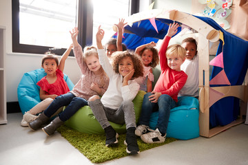 A multi-ethnic group of infant school children sitting on bean bags in a comfortable corner of the classroom, raising their hands to answer a question, low angle, close up