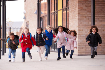 A happy multi-ethnic group of young school kids wearing coats and carrying schoolbags running in a walkway with their classmates outside their infant school building