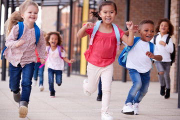 A group of smiling multi-ethnic school kids running in a walkway outside their infant school building after a lesson, close up