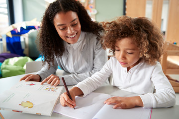Female infant school teacher working one on one with a young schoolgirl sitting at a table writing...