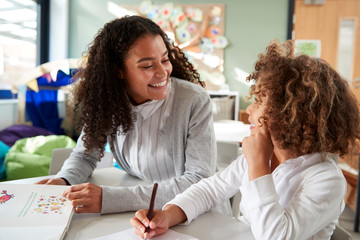 Female infant school teacher working one on one with a young schoolgirl, sitting at a table smiling...