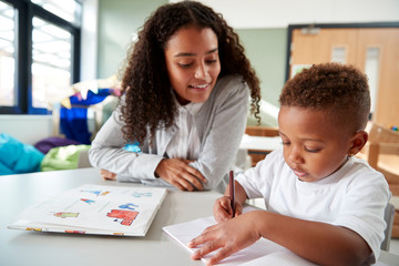 Female infant school teacher working one on one with a young schoolboy, sitting at a table writing...