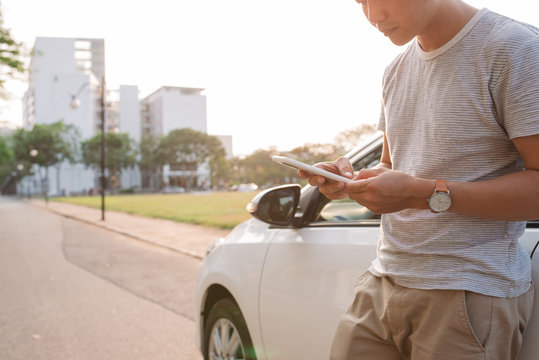 Young Man Is Standing Near The Electric Car And Looks At The Smart Phone. The Rental Car Is Charging At The Charging Station For Electric Vehicles. Car Sharing.