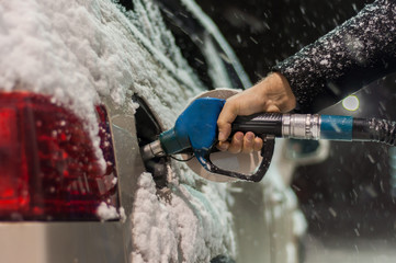 A man in a black coat fills his car at a gas station on a snowy winter evening. Hand holding a blue...