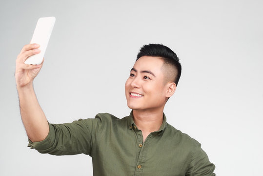 Handsome guy in white shirt is doing selfie using a smart phone, showing Ok sign and smiling, on gray background
