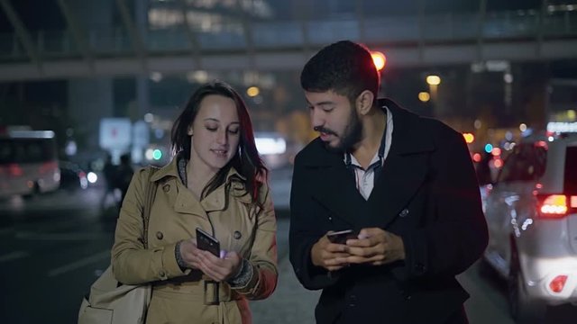Young couple talking and using cell phones on street. Beautiful young woman using smartphone and showing something to smiling handsome man while walking together in night city. Technology concept