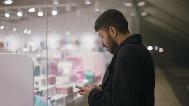 Bearded man messaging by smartphone and looking at showcase. Side view of handsome young man in trench coat using cell phone and standing near shop in the evening. Shopping concept