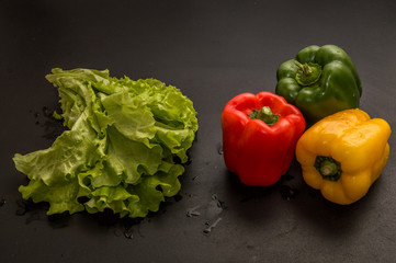 Close up, macro. Flat lay. Three multicolored peppers (paprika) and a big bunch of fresh green lettuce on black background.