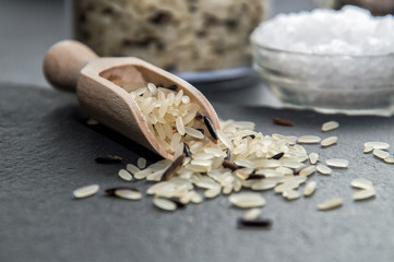 Close up. A wooden spoon full of wild rice. Grains are scattered on a black table, against the...