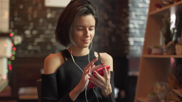 Beautiful girl with short black bob hairstyle, in black shoulderless sweater sitting in cafe alone, listening music, dancing with hair, staring at phone, switching the songs. Blurred background