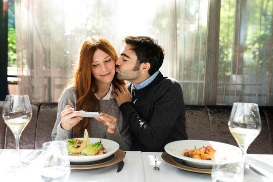 Man kissing woman taking a cell phone picture in a restaurant