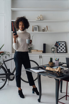 Mid adult freelancer standing in her home office, using smartphone, holding coffee cup