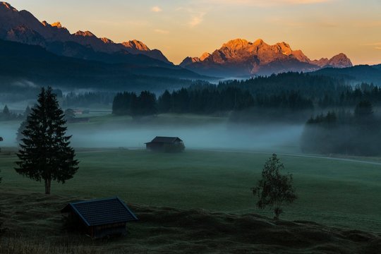 Mountain meadows with small huts in light ground fog and Zugspitz massif in the background at sunrise, Krun, Bavaria, Germany, Europe