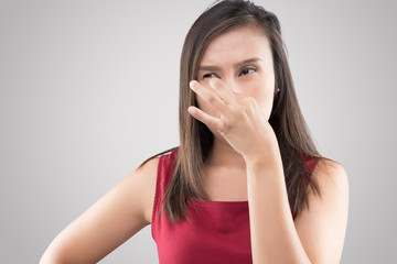 Asian woman in red shirt holding her nose because of a bad smell on gray background.