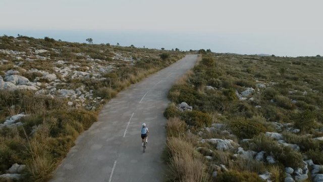 Inspiring and motivational aerial drone video of strong female athlete, achieve her goals, ride bicycle up mountain curvy road at beautiful and epic sunset or sunrise. Concept training or workout