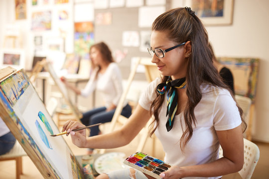 Beautiful brown-haired girl in glasses dressed in white t-shirt and jeans with a scarf around her neck paints a picture in the art studio