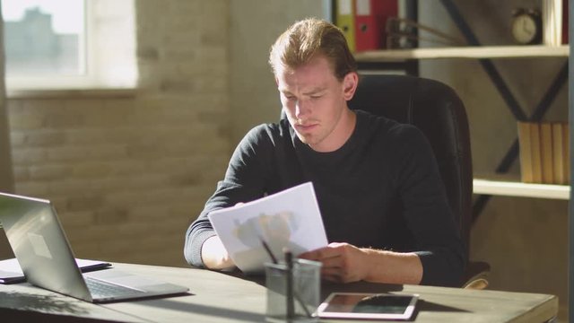 Young concentrated businessman sitting in the office at the table near notebook and working at documents, make amendment in papers.