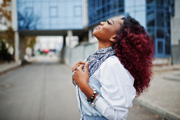 Attractive curly african american woman in jeans dress posed against modern multistory building.