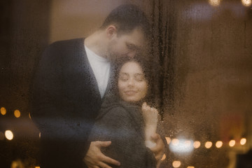 Loving guy hugs and kisses his girlfriend standing behind a wet window with lights. Romantic couple