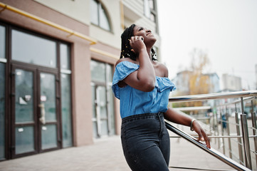 Fototapeta premium Attractive african american woman with dreads in jeans wear posed near railings against modern multistory building speaking at mobile phone.