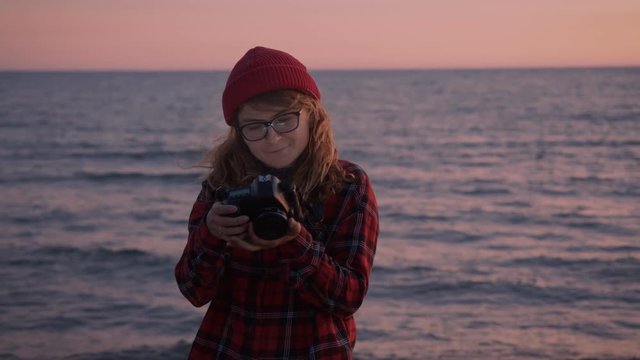 Romantic shot of young woman or millennial teenager with professional photo camera stand on sunset beach in soft pink colors. Looks in lens and smiles gently.Creates images for social media influencer