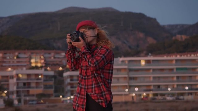 Cute hipster young woman in red outfit stands on sunset beach, looks and smiles into camera. Starts making photographs for memories or content creation. Influencer creative job