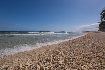 beach with coral stones pebbles at the seashore with waves and boats in the background on a day with blue sky and white clouds  