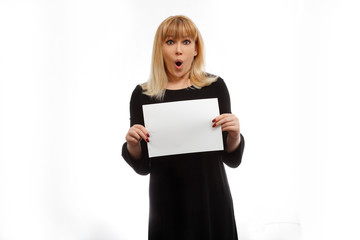 Fototapeta premium Surprised blonde model in a black dress with a sheet for the text in the hands on a white background. Emotions model. Surprise and invitation. Place for text. A model mouth open. Strong surprise