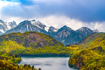 Naklejka na ściany i meble Great panoramic view of the lake Alpsee, the surrounding hills with forest & the amazing snow-capped mountain tops of the Alps in the background. The beautiful nature is a popular tourist destination.