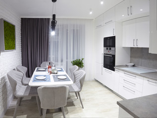 White kitchen interior with grey chairs and served table with white plates glasses and moss on the wall in new luxury home with lights on.