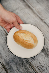 Man's hand holding plate with German style bread roll or bun, so called Schrippe or Brötchen, a small loaf made of wheat flour traditionally served for breakfast on white plate and wooden background