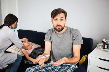 Distracted young man hold thermometer and look on camera. He is frustrated. Doctor sit besides sick girl. She lying on sofa.