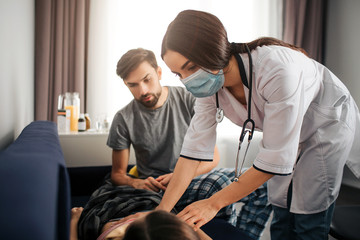 Young female doctor stand upon sick girl. She look at her and touch. Father sit besides. He looks worried.