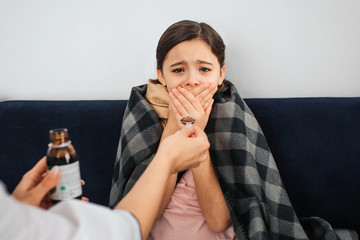 Small girl look on camera and cover mouth with hands. She is frightened. Female doctor give her...