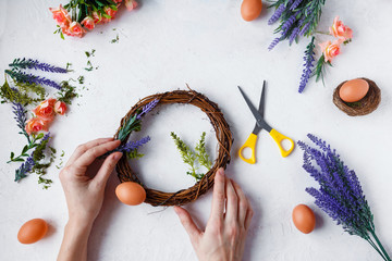 Female hands make Easter wreath of flowers, herbs and eggs on a light background. Easter concept