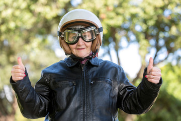 Motociclista anziana con casco occhiali e giubbotto in pelle fa l'okay con i pollici - sfondo natura