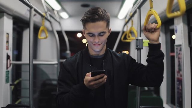 Portrait of attractive men holds the handrail, using smartphone standing in public transport. City lights background