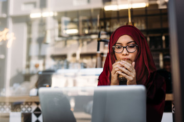 Woman in hijab using laptop and having coffee at cafe