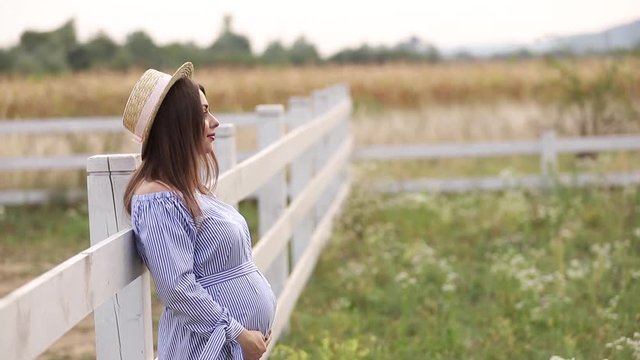Pregnant woman in nature try on knitted hat. Background of field and white fance near the farm
