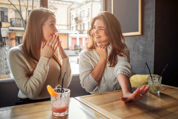 Cheeful and happy young women sit at table. First show hand. Another model wonderes. They are happy.