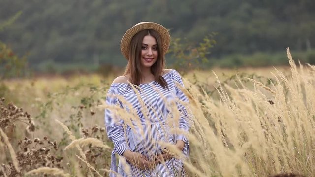 Pregnant woman in nature try on knitted hat. Background of field and white fance near the farm. mountains