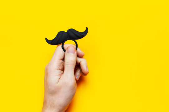 Men's hands hold Black mustache, props for photo booths, carnival, parties on yellow background top view flat lay copy space Father's day Creative party decoration concept