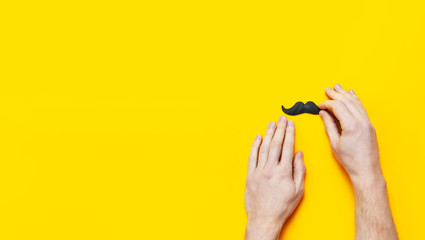 Men's hands hold Black mustache, props for photo booths, carnival, parties on yellow background top view flat lay copy space Father's day Creative party decoration concept