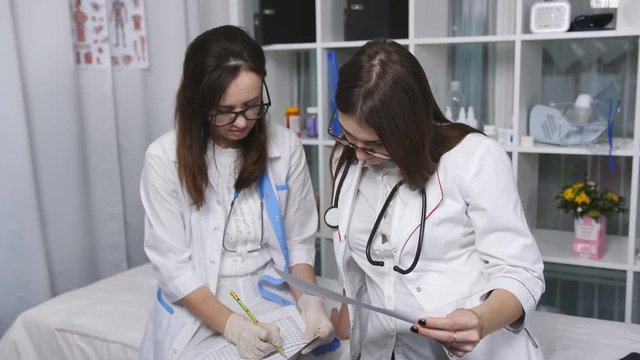 Two beautiful young doctors women double check files with paper documents. Two women doctor in the examining room filing papers.