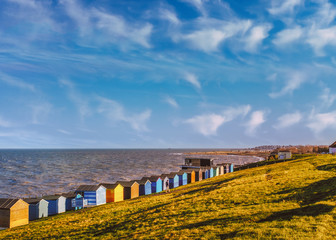 Row of beach huts along the coast in Tankerton, Whitstable, Kent. The green grass slopes are behind the huts and a man strolling along the grass. The clouds in the blue sky are soft and whispy