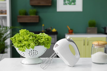Colander with fresh lettuce, mixer and jar with flour on table in kitchen
