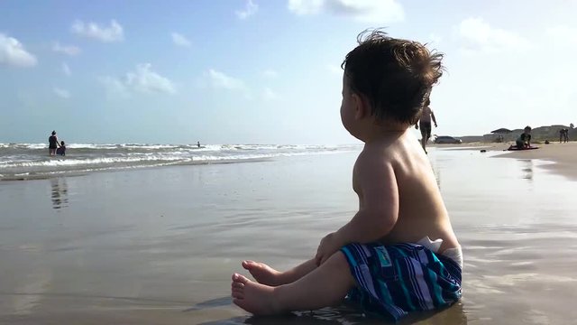 Toddler reaching out to the ocean sitting on the shore.