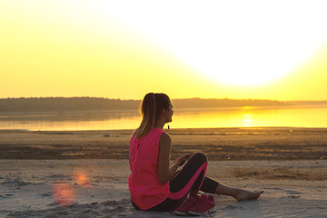 Fototapeta na wymiar Young beautiful girl in sportswear sits on a sandy beach and enjoys an orange sunset by the sea and listens to music in her mobile phone